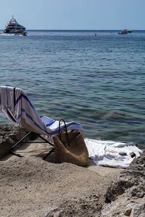 Una silla de playa con una toalla a rayas y un bolso tejido sobre una costa rocosa, con vista a un mar tranquilo donde se ven barcos a lo lejos.