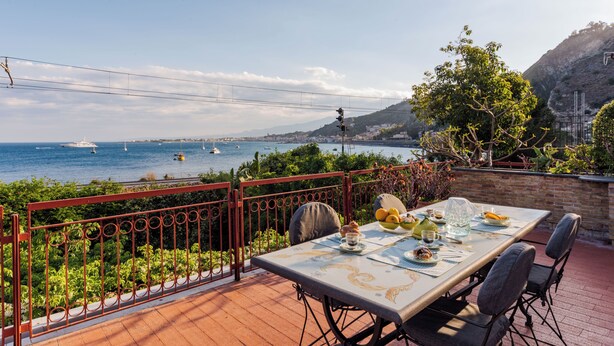 A terrace with a table set for dining, overlooking a coastal area with boats and hills. - TámuTaormina | Luxury Sea & Etna Views Duplex (Taormina)