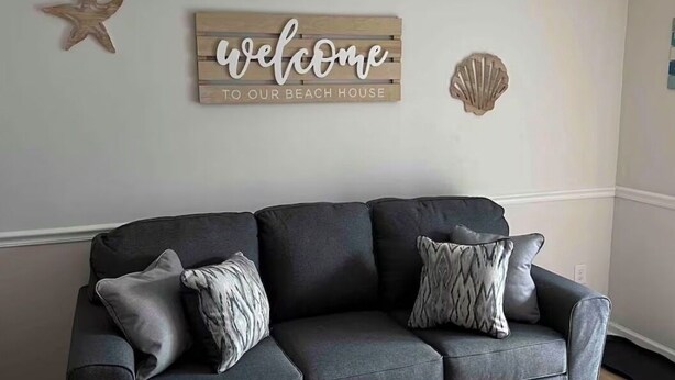 Um sofá cinza escuro com almofadas estampadas, uma placa de madeira com a inscrição 'welcome to our beach house', uma decoração de estrela-do-mar e uma concha na parede.