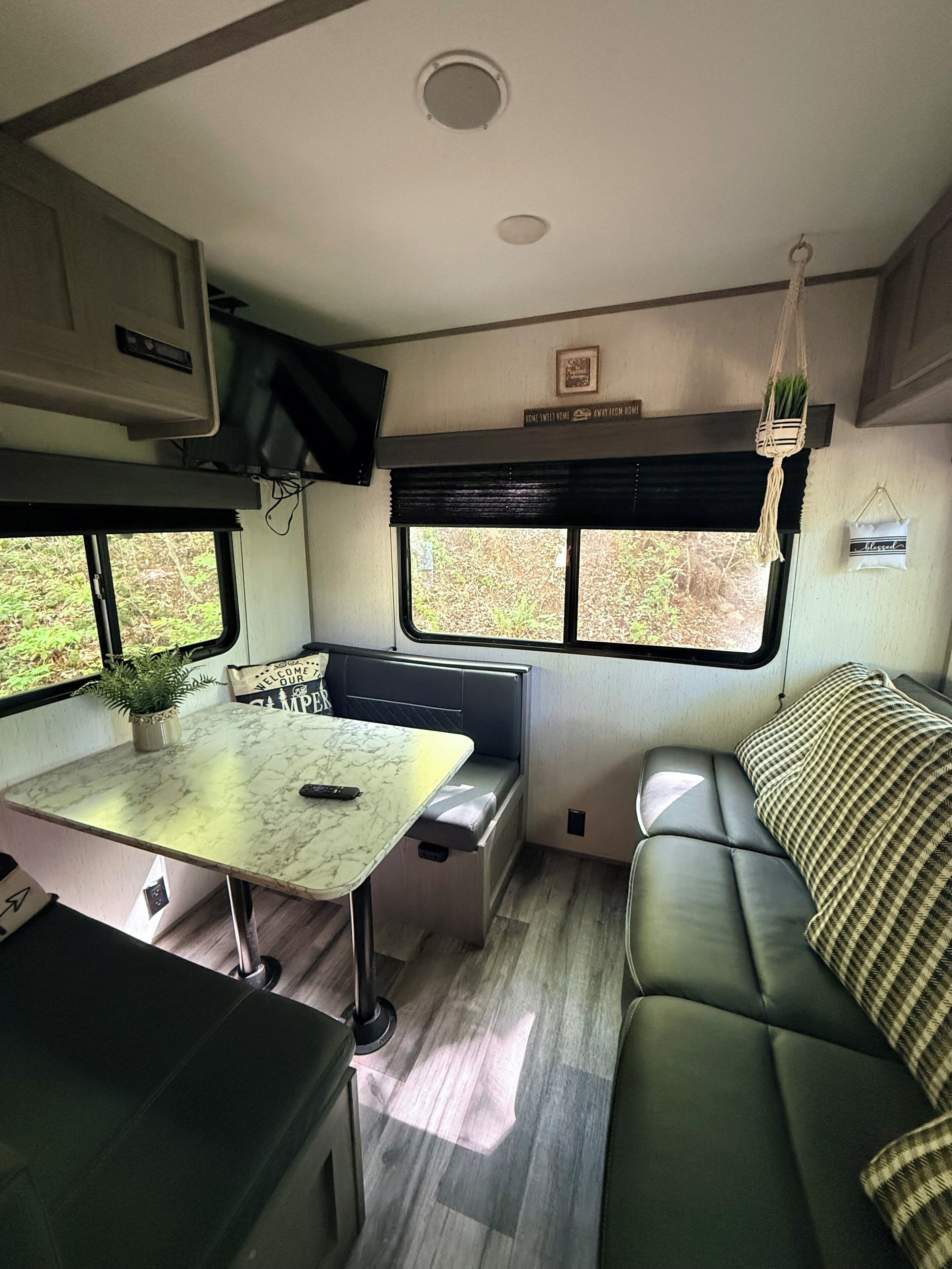 A compact RV interior with a dining area, seating, a mounted TV, and a window with a view of greenery.