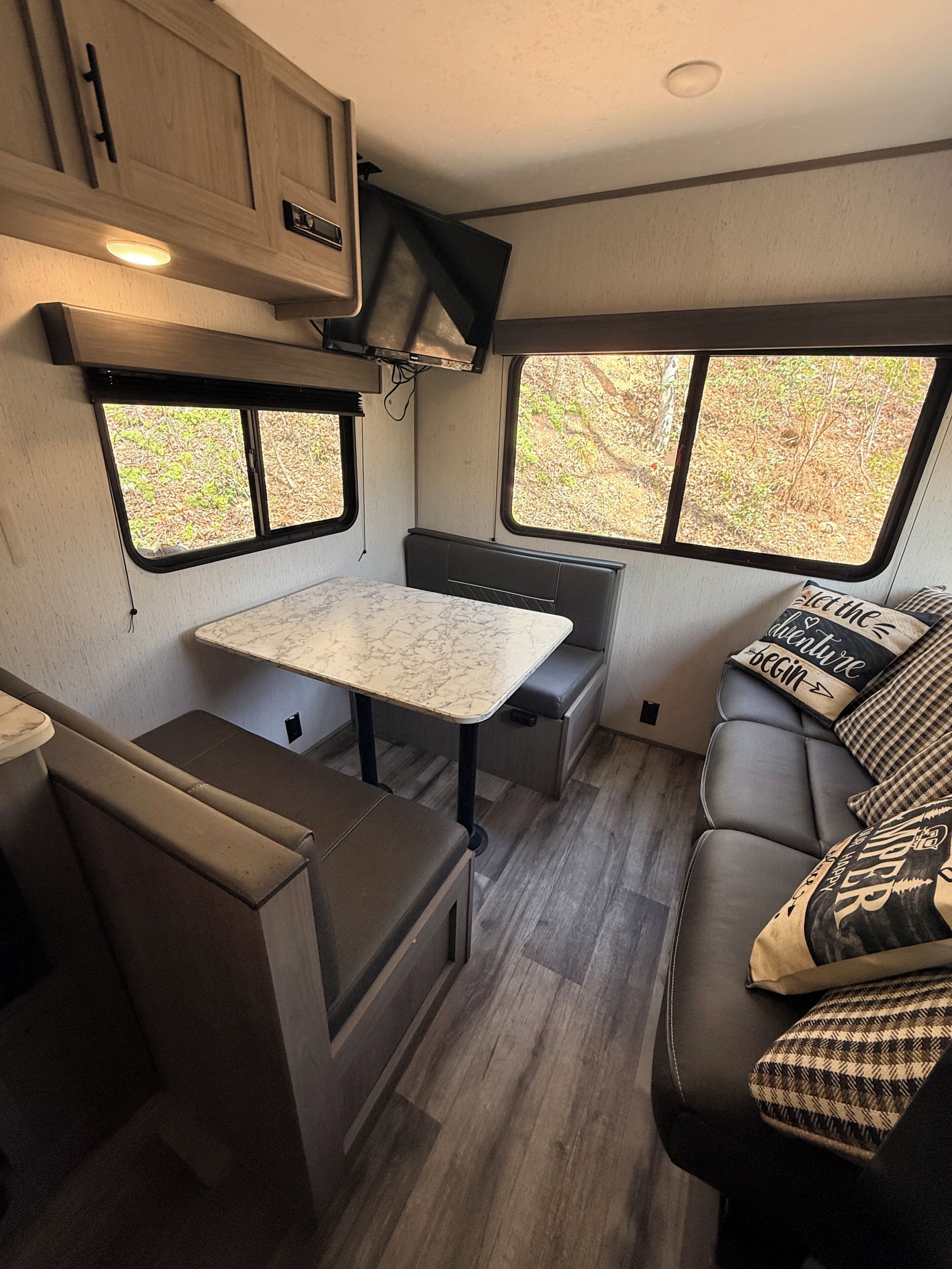 A compact RV interior with a dining table, seating area, and windows providing views of the outdoors.