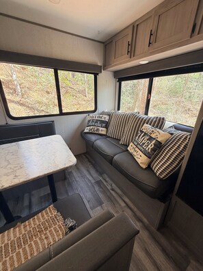 An RV interior with a dining table, a sofa with decorative pillows, and windows offering a view of trees.