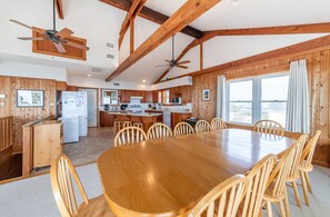 A spacious kitchen with wooden beams, a dining area with a large wooden table, and a view through large windows. - Anna - Corolla, Ocean Sands, Oceanfront, 4 Bedrooms, Ocean Views, Private Walkway to the Beach (Corolla)