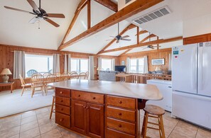 A spacious kitchen with wooden beams, a central island with drawers, a refrigerator, and a dining area with chairs and a table. - Anna - Corolla, Ocean Sands, Oceanfront, 4 Bedrooms, Ocean Views, Private Walkway to the Beach (Corolla)