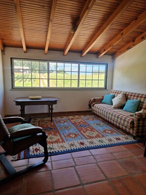 A room with a wooden ceiling, a large window with a view of a field, a plaid sofa, a patterned rug, and a wooden chair.
