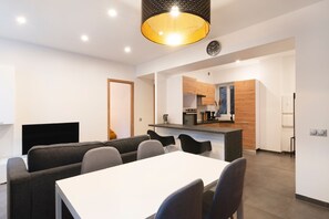 Une cuisine moderne avec des meubles en bois et un îlot central, un coin repas avec une table blanche et des chaises grises, et un grand téléviseur à écran plat fixé au mur.