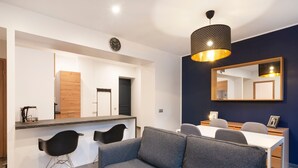 Een moderne woonkamer met een bank, een eettafel en stoelen. Er is een keukenhoek met een koffiezetapparaat en een klok aan de muur.