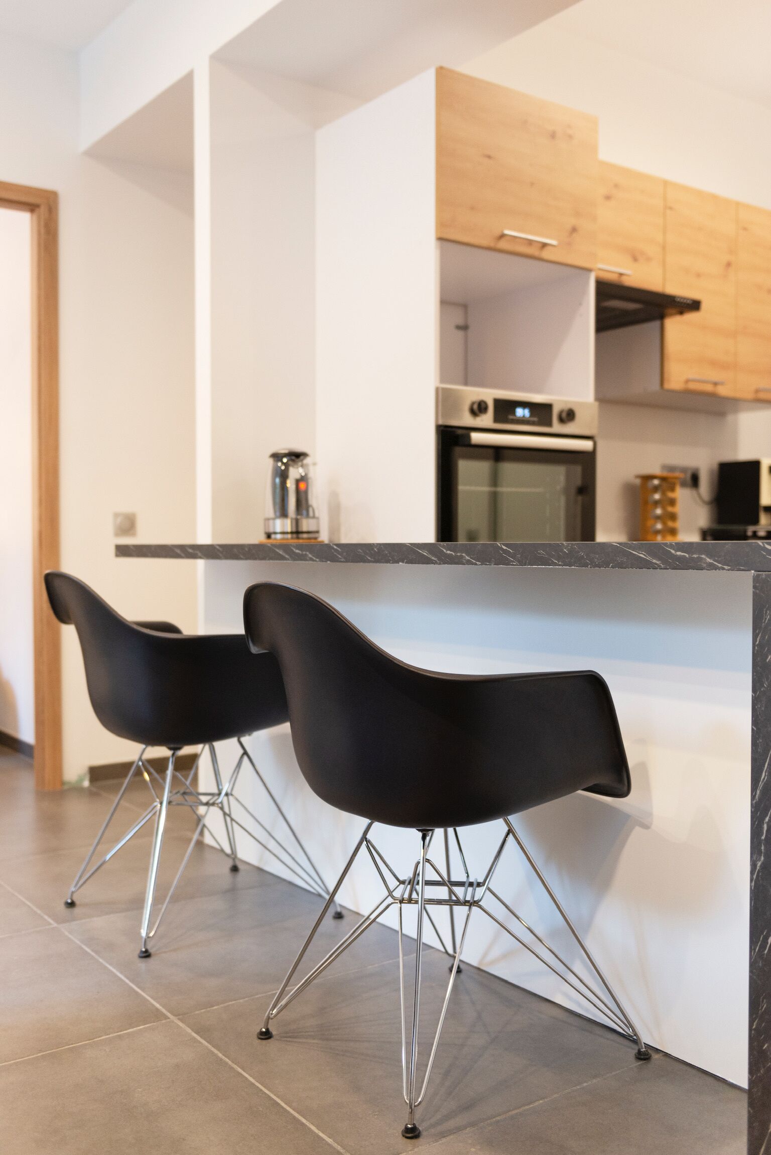 Une cuisine moderne avec deux tabourets de bar noirs, un comptoir en marbre, des appareils électroménagers en acier inoxydable et des armoires en bois.
