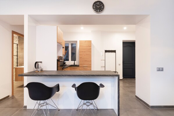 Une cuisine moderne avec un plan de travail en marbre, deux tabourets de bar noirs et une horloge au mur.