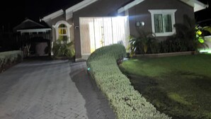 Una casa de dos pisos con entrada pavimentada, césped cuidado y un ventanal con cortinas.