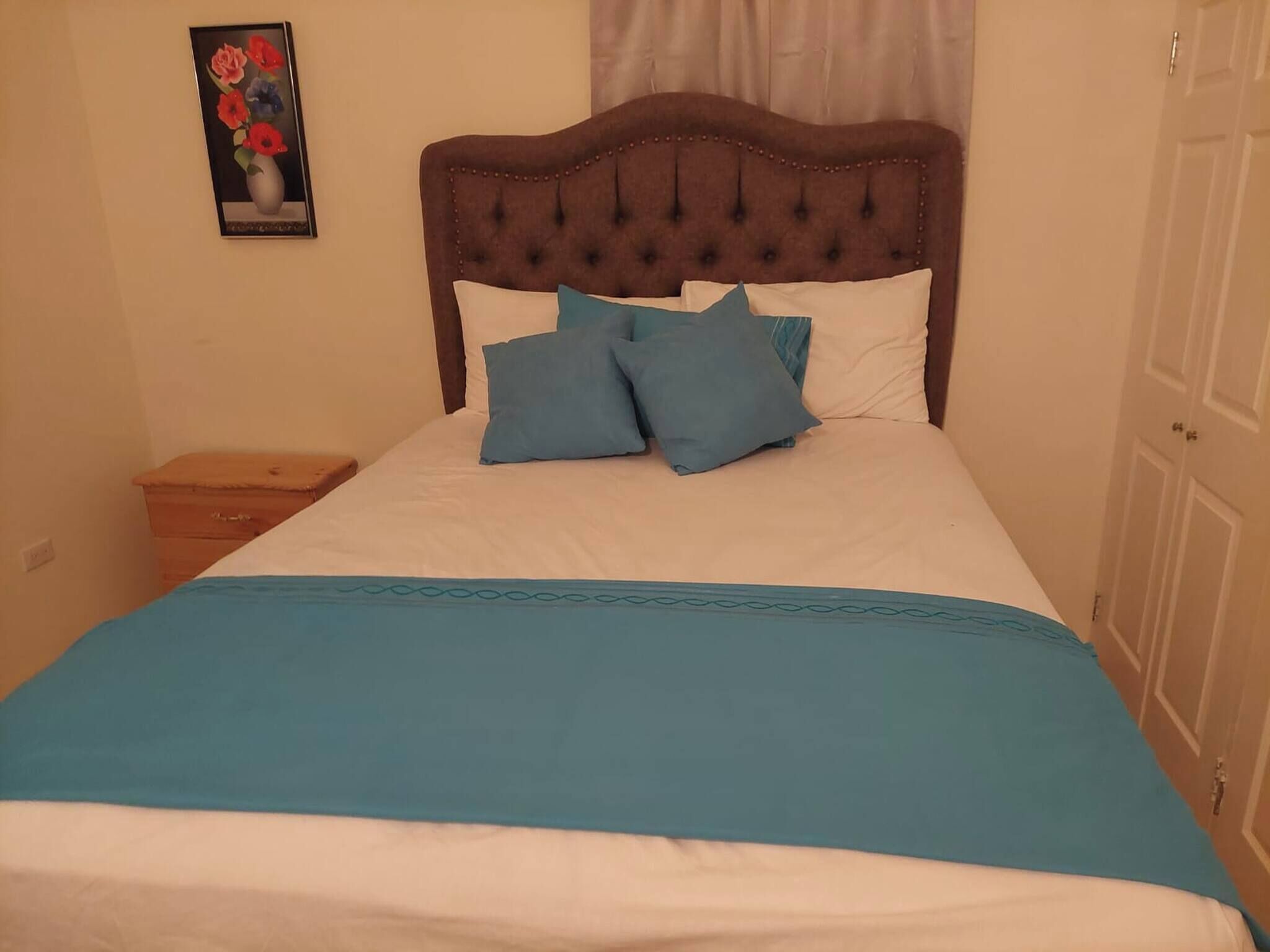 Una cama con cabecera capitonada, dos almohadas, una colcha azul y una mesita de noche con un cajón.