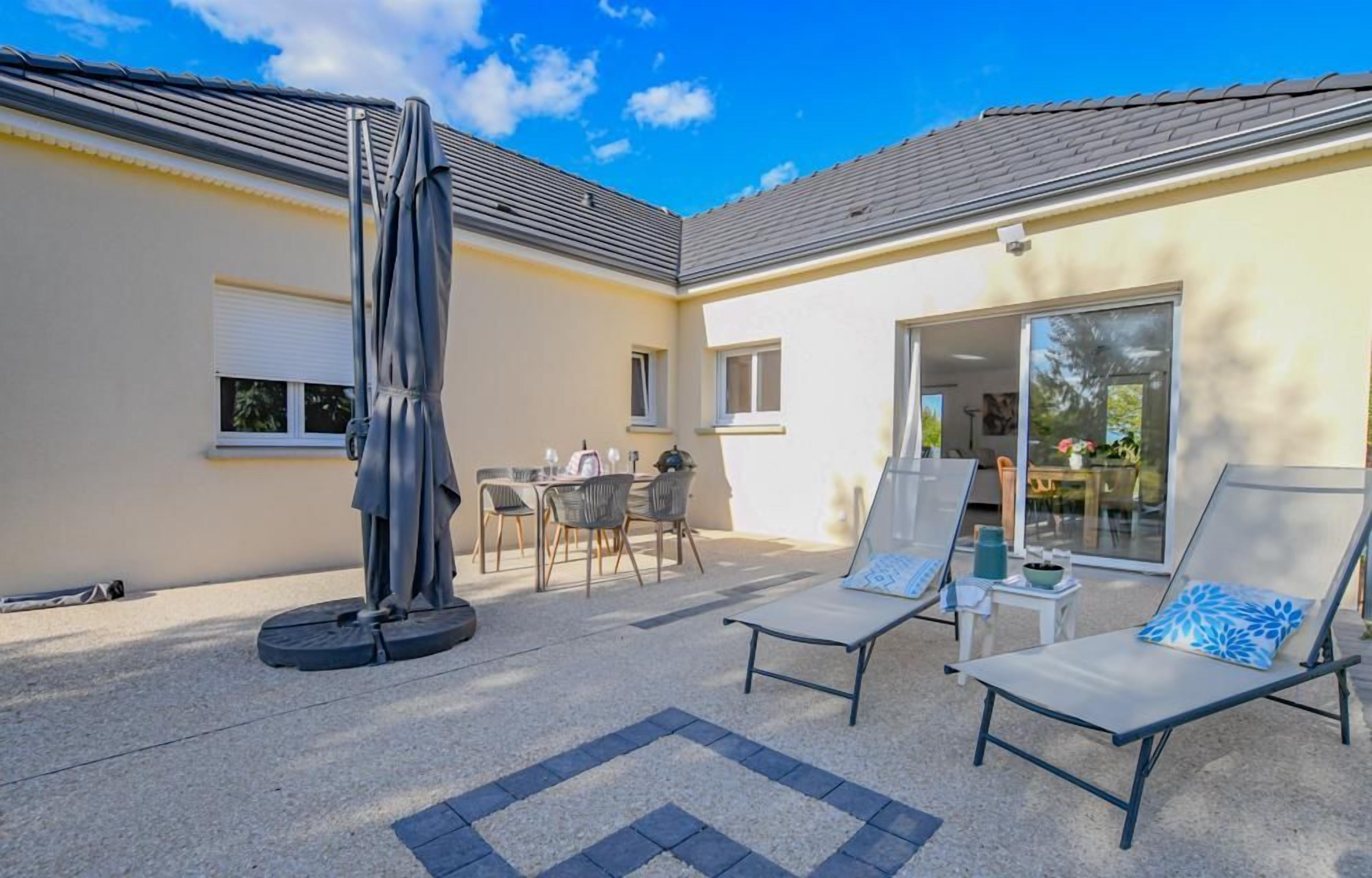 Un espace extérieur abrité, équipé d’une table et de chaises, d’un parasol et de deux fauteuils de jardin avec coussins.