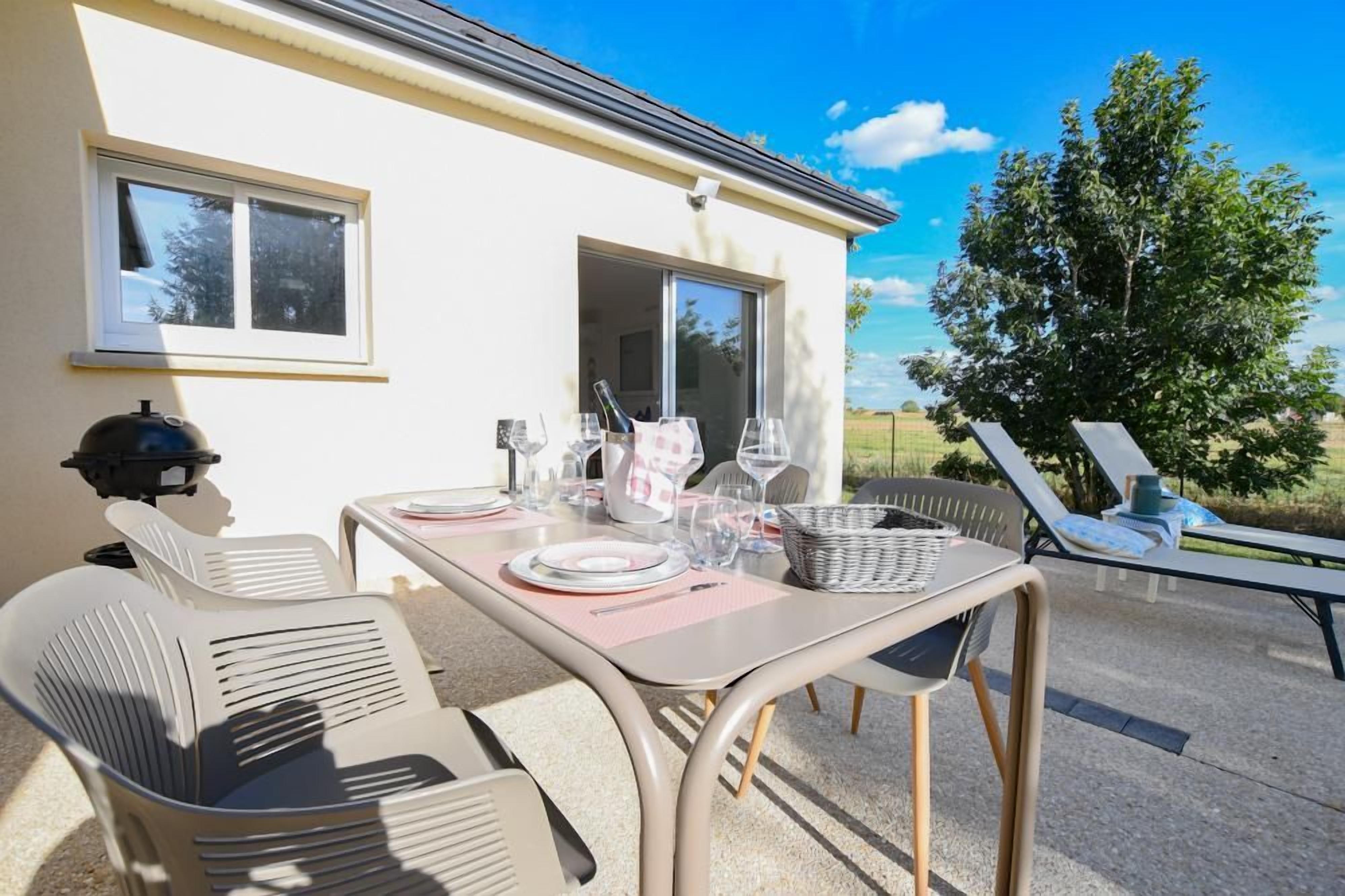 Un espace extérieur aménagé pour prendre les repas, avec une table dressée pour deux personnes, des chaises et une vue sur la campagne.