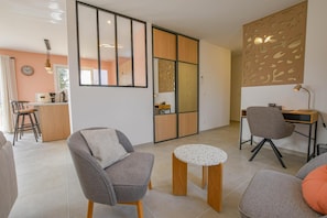 Ein modernes Wohnzimmer mit einer Couch, einem kleinen runden Tisch, einem Schreibtisch mit Stuhl und einer Küchenzeile im Hintergrund.