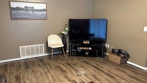 Un salon avec un téléviseur à écran plat, un fauteuil beige, un vase décoratif et un plancher en bois.