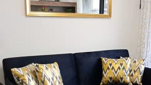 Un sofá azul oscuro con cojines estampados en dorado y negro, un espejo con marco dorado arriba, y suelo de madera gris claro.