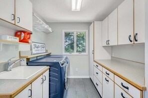 A laundry room with a washing machine, a sink, and a window with a view of trees.