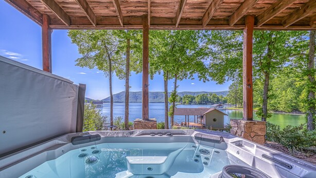 Un bain à remous surmonté d’une pergola en bois, offrant une vue sur un lac bordé d’arbres et sur un bâtiment situé au loin.