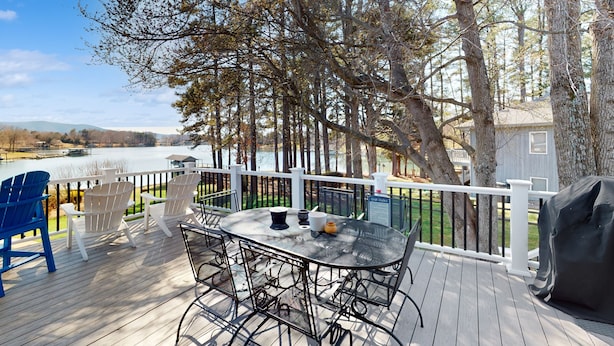 Une terrasse avec une table et des chaises donnant sur un lac, entourée d’arbres et d’une clôture blanche.