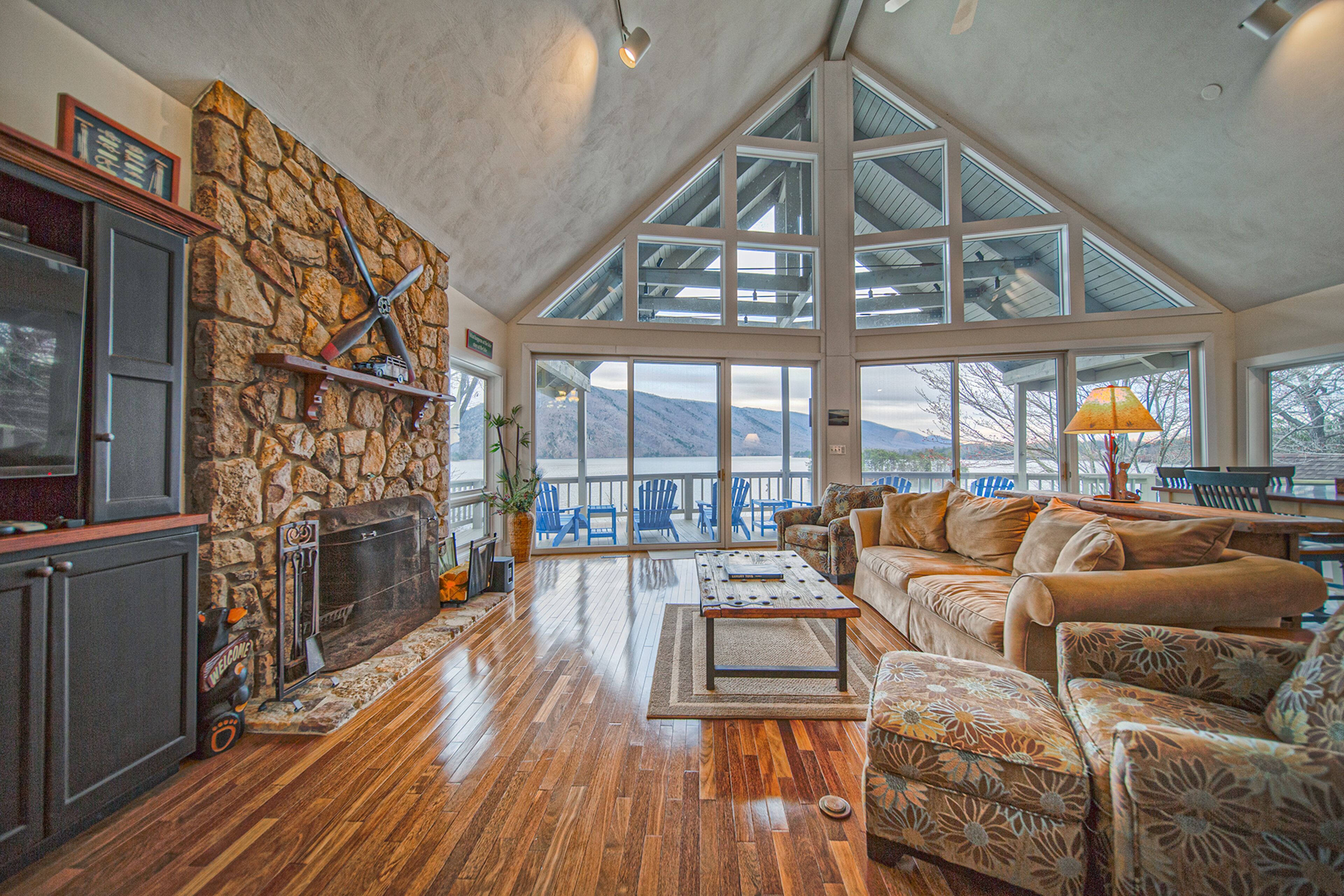 A spacious living area with a stone fireplace, wooden flooring, and large windows offering a view of the outdoors.