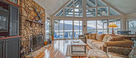 A spacious living area with a stone fireplace, wooden flooring, and large windows offering a view of the outdoors.