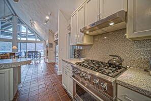 A kitchen with a large stove, granite countertops, and white cabinets. There is a dining area with blue chairs and a table visible through large windows. - Our Point of View - Lakefront home with hot tub, pool table, dock - dog-friendly (Huddleston)