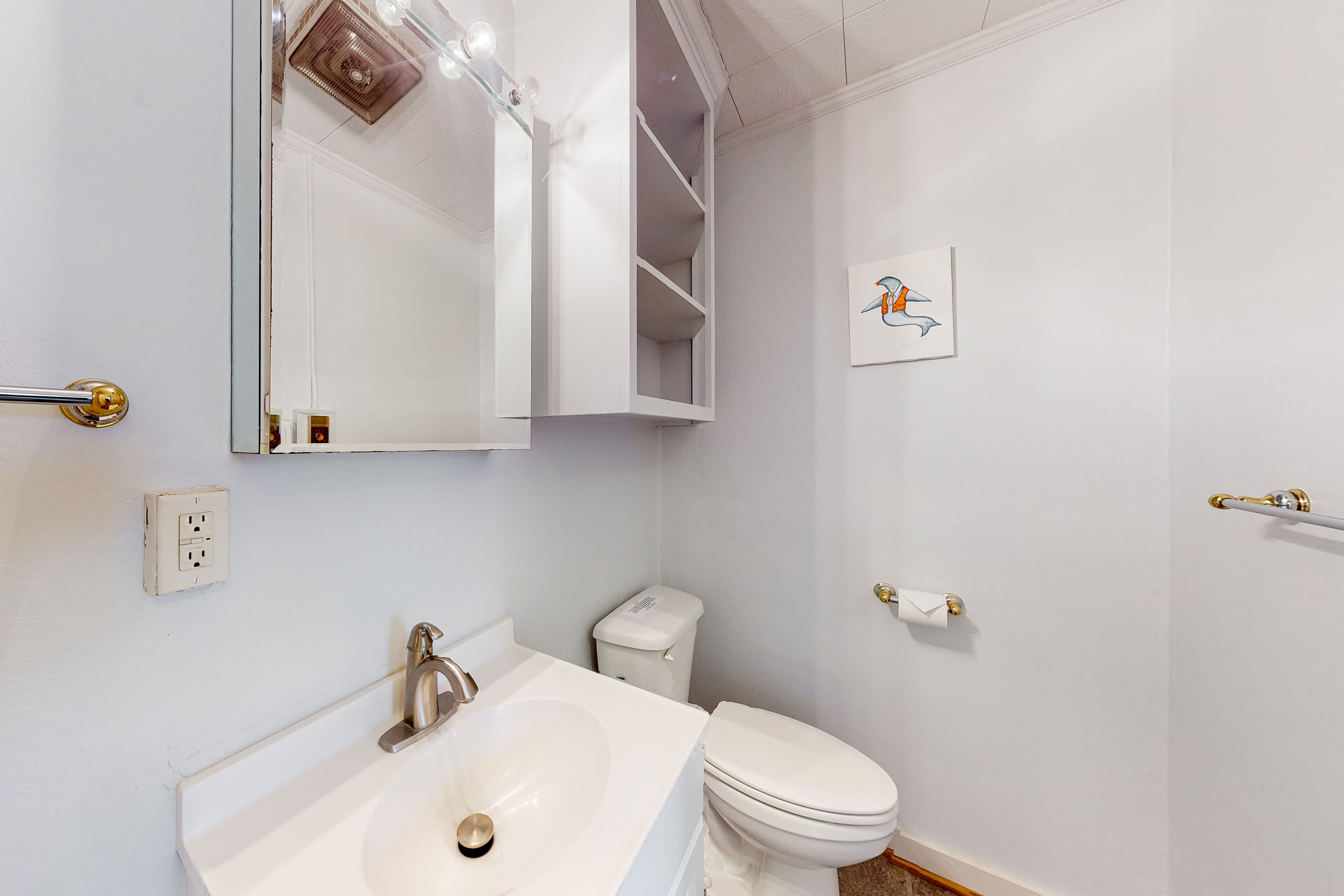 Une salle de bain avec un lavabo blanc, des toilettes et un miroir au-dessus du lavabo. Il y a une étagère au-dessus des toilettes et un luminaire au-dessus du miroir.
