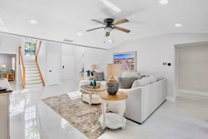 A spacious living room with a white sofa, a coffee table, and a ceiling fan. A staircase is visible in the background leading to another room. - NEW! Heron Lagoon Hideaway: Canal Front, Pool, Dog (Sarasota)