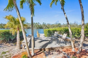 Une terrasse en bois, agréée de mobilier d’extérieur, entourée de palmiers et bordée par une étendue d’eau.
