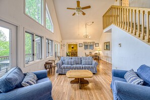 A spacious living room with a high ceiling, a wooden ceiling fan, a blue sofa set, a wooden coffee table, and a staircase with wooden railings.