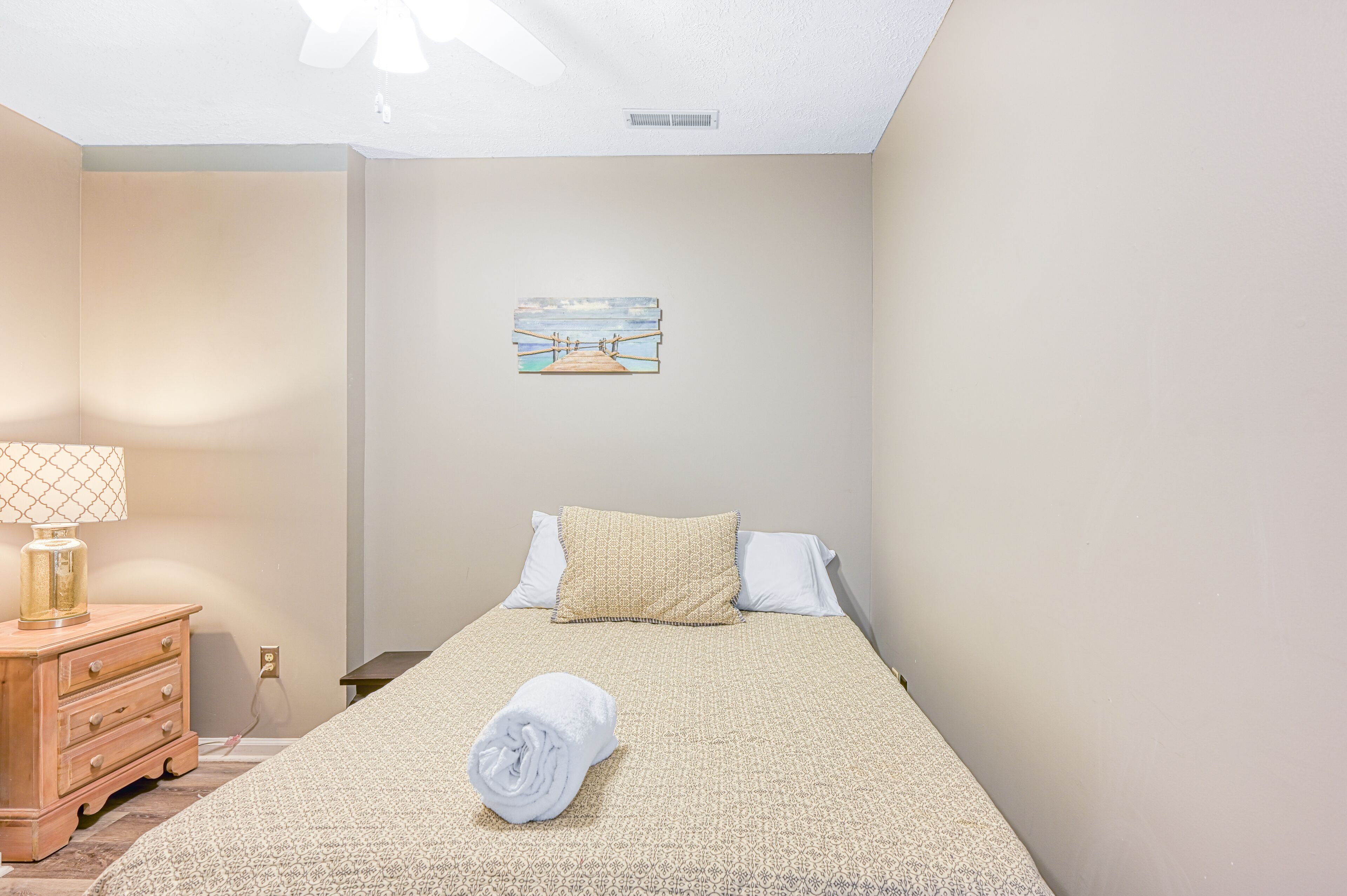 A neatly made bed with a patterned bedspread, a rolled white towel, and a painting on the wall.