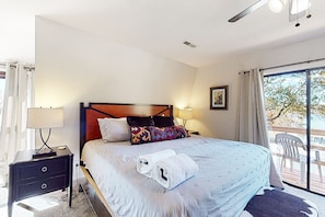 Un lit bien fait, recouvert d’une couvre-lit à motifs, avec une tête de lit en bois et deux oreillers. On aperçoit une table de chevet avec une lampe et un petit tiroir. Une fenêtre avec des rideaux est visible, et une chaise se trouve à l’extérieur, devant la fenêtre.
