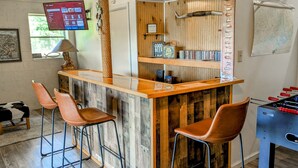 A bar area with stools, a foosball table, and a TV screen displaying a menu. - Dock Sun Point - Lakefront home for easy boating, fishing, private dock & AC/Wifi, Dogs ok! (Huddleston)