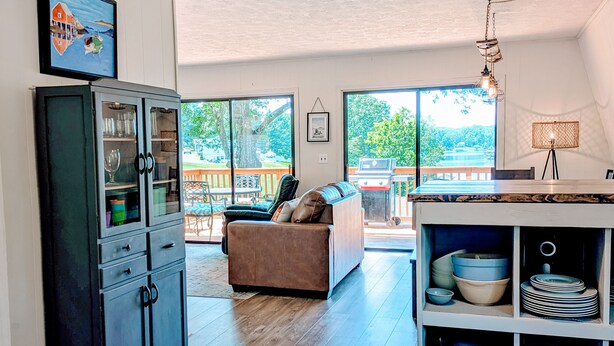 A spacious living area with a large window, a sofa, a cabinet, and a dining area with a table and chairs. - Dock Sun Point - Lakefront home for easy boating, fishing, private dock & AC/Wifi, Dogs ok! (Huddleston)