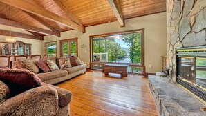 A spacious living room with wooden floors, a stone fireplace, and large windows offering a view of greenery. - The Main House - Lakefront home with private hot tub & balcony (Huddleston)