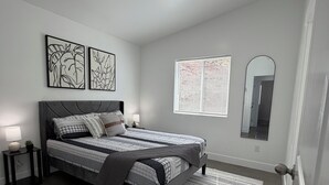 Un dormitorio moderno con cama, mesitas de noche, ventana con persianas, espejo y obra de arte en la pared.
