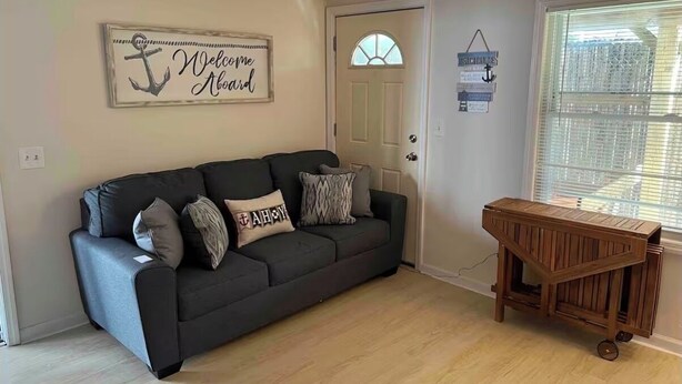 Un salon avec un canapé gris foncé, une table d’appoint en bois, une porte avec une fenêtre et un panneau mural sur lequel on peut lire : « Bienvenue à bord ».
