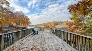 A wooden deck with a chair, overlooking a lake with autumn foliage and houses in the background.