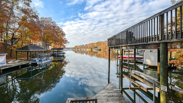 A calm river with boats docked on the left, a wooden pier on the right, and autumn trees reflecting in the water.