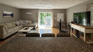 A living room with a large TV, a sofa, a coffee table, and a dining area with a granite countertop. - Humble Hideaway - Inviting lakefront home with firepit, balcony & W/D - dog-friendly (Moneta)