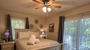 A bedroom with a wooden bed, two bedside tables with lamps, a ceiling fan, and a large window with curtains. - Humble Hideaway - Inviting lakefront home with firepit, balcony & W/D - dog-friendly (Moneta)
