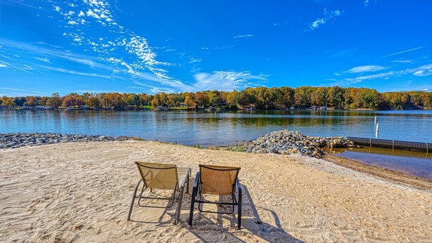 Une plage avec deux chaises donnant sur un lac, entourée d’arbres aux couleurs automnales et sous un ciel dégagé.