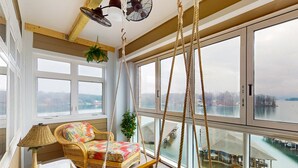 Une pièce spacieuse avec de grandes fenêtres donnant sur un plan d’eau, un canapé en osier, une balançoire suspendue et une lampe posée sur une petite table.