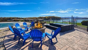 Une terrasse avec des fauteuils Adirondack bleus, un foyer et un jacuzzi donnant sur un lac.