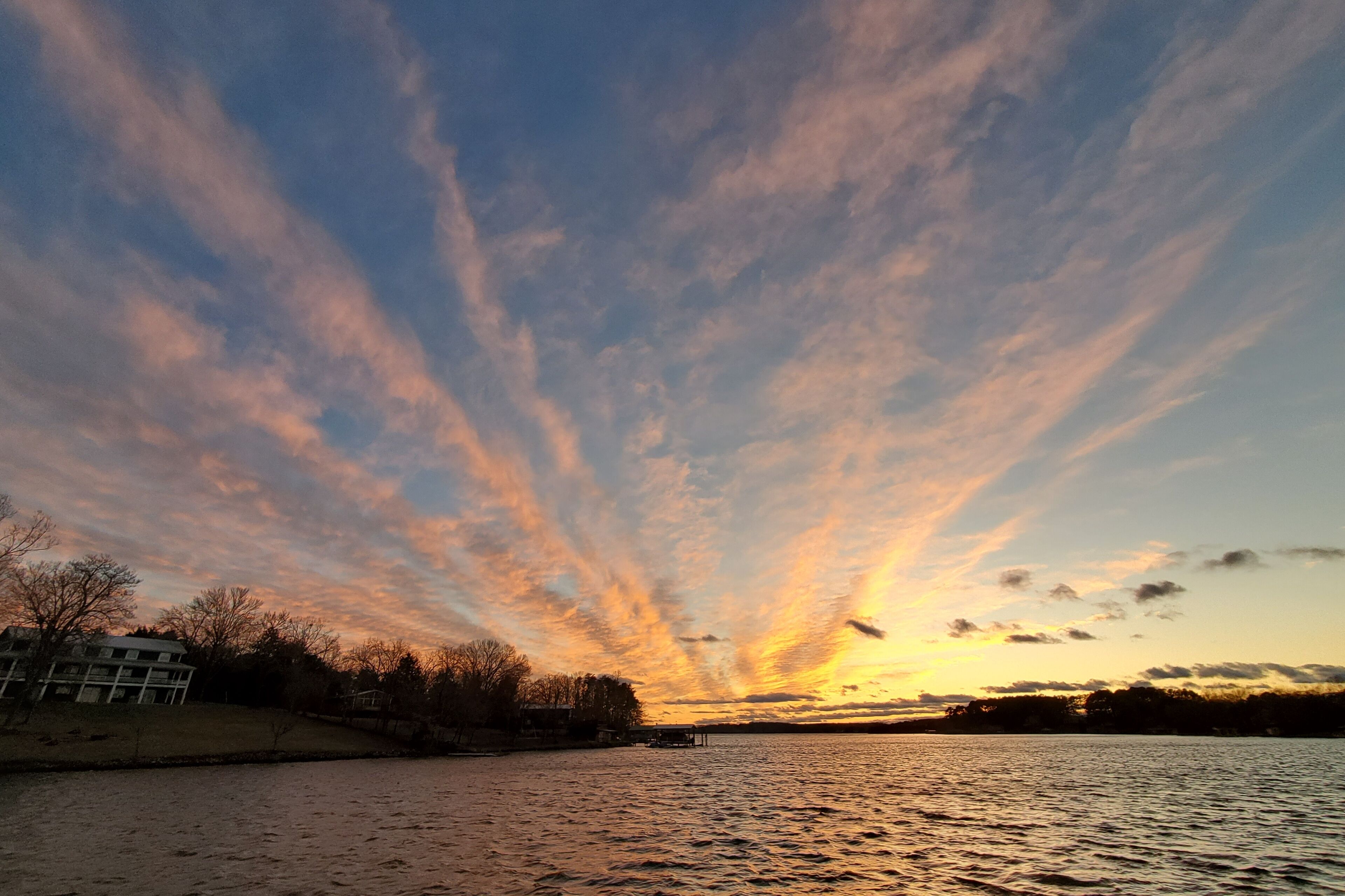 A sunset over a body of water with a building on the left and trees in the foreground.