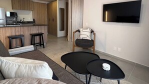 Ein modernes Wohnzimmer mit einer Couch, zwei schwarzen Tischen, einem an der Wand befestigten Fernseher und einer Küchenzeile im Hintergrund.