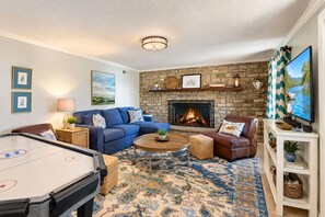 A cozy living room with a pool table, a blue sofa, a leather armchair, a fireplace, and a television. - Smith Mountain Bliss - Spacious waterfront house w/patio, deck, fire pit & grill (Moneta)