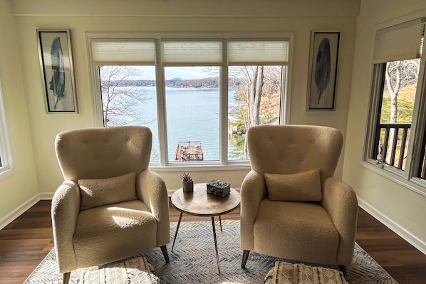 A room with two beige armchairs facing a window overlooking a lake, a small round table with a potted plant, and a patterned rug. - LakeDaze - Dog-friendly, Lakefront, Dock, Hot Tub, Putting Green, Game Room (Moneta)