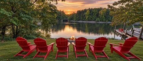 A lakeside view with a row of red Adirondack chairs facing a campfire, surrounded by trees and a dock.