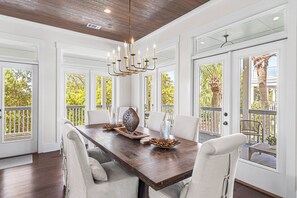 A dining area with a wooden table, white chairs, a chandelier, and large windows with a view of trees. - 392 Walton Rose - Access to Rosemary Beach Fitness, Tennis, 4 Adult bikes! (Inlet Beach)